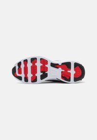 Semelle d'une chaussure de sport avec une base blanche, des motifs de bande de roulement en caoutchouc rouge et noir et un petit logo swoosh noir près de la zone du talon.