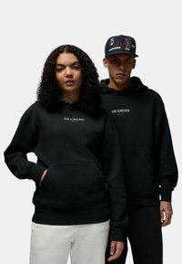Le felpe nere hanno "AIR JORDAN" ricamato sul petto. Il design presenta una tasca a marsupio e polsini a coste. Il modello a destra indossa un cappellino abbinato.