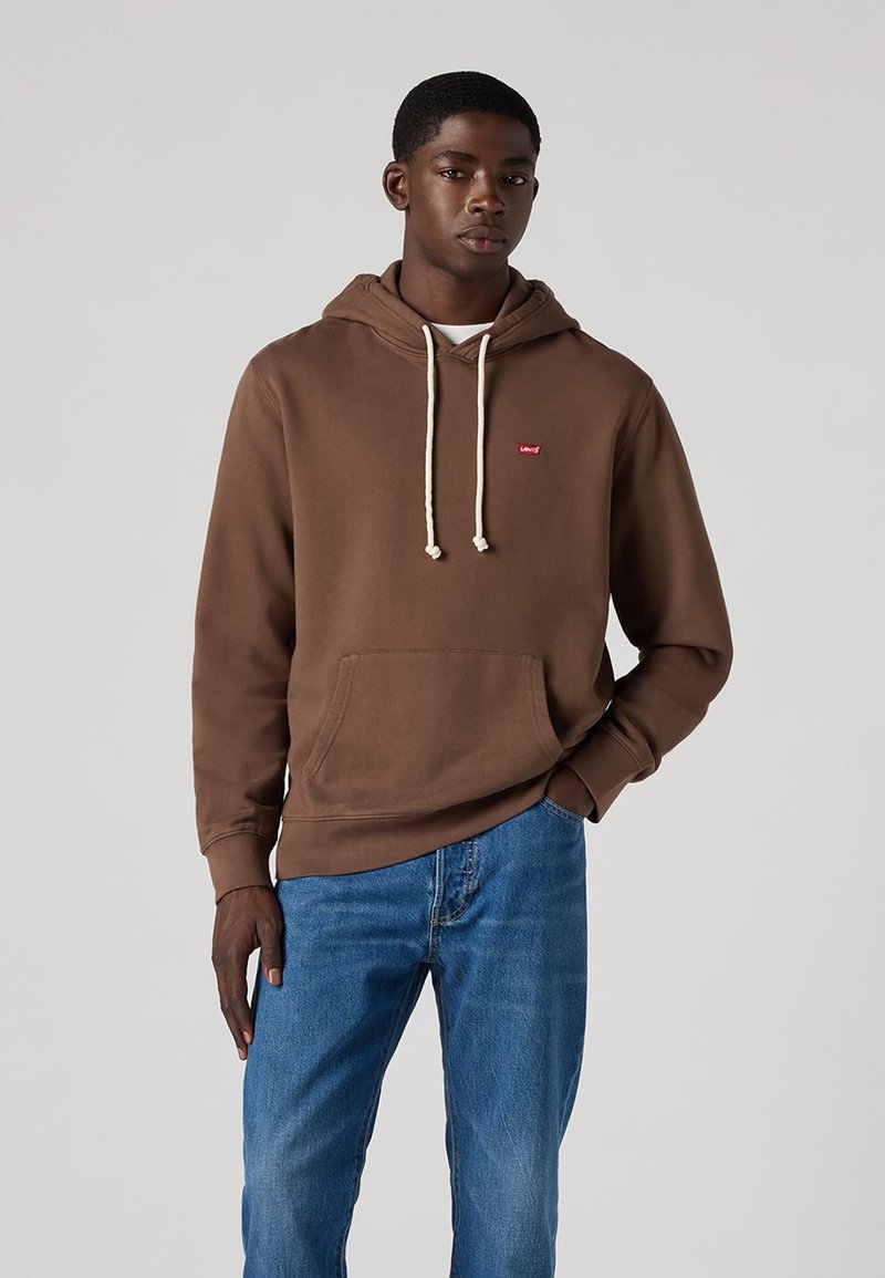 Felpa marrone con tasca a marsupio, cappuccio regolabile con lacci e piccolo logo rosso. Indossata con jeans blu dal taglio classico.