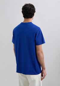 Uomo con capelli neri ricci che indossa una camicia blu a maniche corte semplice e pantaloni di un colore chiaro, in piedi di fronte a una parete grigia chiara e uniforme.