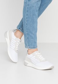 Białe i szare buty sportowe mają teksturowaną powierzchnię, sznurowany design, subtelny metaliczny akcent i wyściełaną podeszwę dla wsparcia.