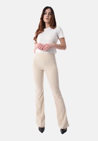 Donna in pantaloni a campana beige, top bianco a maniche corte e scarpe nere a punta, davanti a uno sfondo chiaro e uniforme.