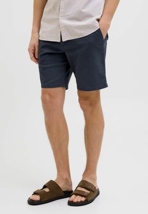 Hombre con pantalones cortos azul marino, camisa beige abotonada y sandalias marrones de punta abierta, de pie con una mano en el bolsillo.