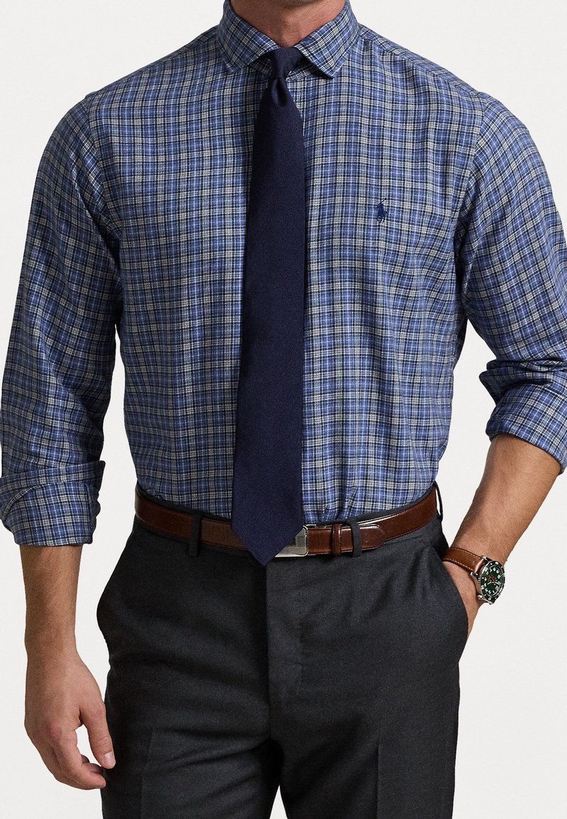 Blå och vit rutad skjorta med lång ärm, knäppta manschetter och ett marinblått slips. Buren med grå byxor och ett brunt bälte.