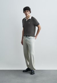 Polo marron avec col blanc, jean en denim clair, ceinture noire et chaussures noires ; le modèle se tient devant un fond neutre, mains détendues.