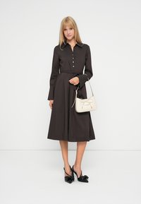 Langärmliges braunes Kleid mit einem geknöpften Oberteil und einem taillierten Gürtel, kombiniert mit einer cremefarbenen Handtasche und schwarzen High Heels. Glatte Textur.