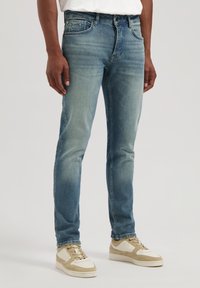 Jeanși din denim albastru deschis, cu o croială slim, având decolorări subtile și detalii de tip whiskering. Împărțiți cu adidași bej, prezentați din talie până la picioare.