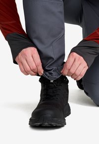 Händer justerar ärmen på grå byxor över svarta vandringsskor, person klädd i en röd och grå långärmad jacka, knäböjande på ett knä.