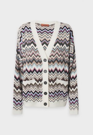 Un mannequin se tient devant un fond uni, portant un cardigan multicolore à motifs en zigzag, un haut blanc, une ceinture marron et un jean gris à jambes larges.