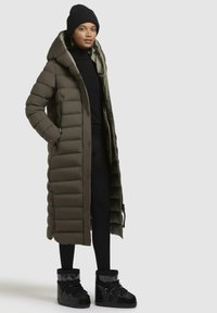 Padded long coat in olive green with a hood, featuring a button front and side pockets. Paired with black ankle boots and leggings.