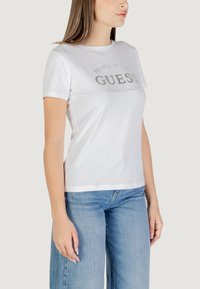 T-shirt bianca in cotone con maniche corte, caratterizzata dal logo in argento metallico "GUESS" e dall'anno di fondazione, abbinata a jeans blu sbiaditi.