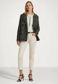 Mulher a usar um blazer de tweed preto e branco, uma camisa branca com riscas pretas, calças bege e sandálias de salto alto pretas, em pé com uma mão no bolso.