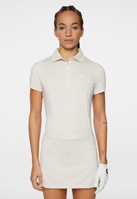 Beige polo t-shirt med korta ärmar och en tvåknappslucka, parat med en matchande kjol. Har en vit logotyp på t-shirten.