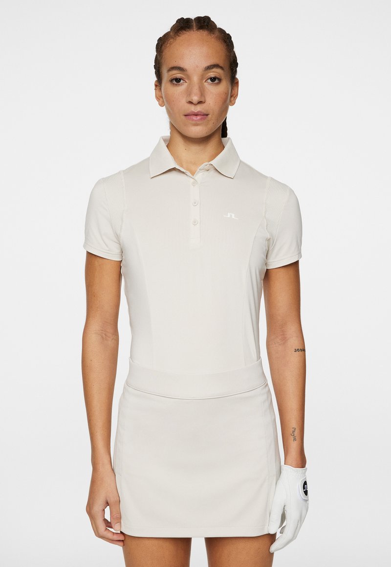 Beige polo t-shirt med korta ärmar och en tvåknappslucka, parat med en matchande kjol. Har en vit logotyp på t-shirten.