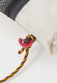 Beige Tasche mit strukturierten Oberflächen, ausgestattet mit einem roten Kunststoffverschluss und einem geflochtenen, mehrfarbigen Zugband für sicheren Verschluss.