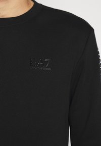 Black long-sleeve shirt made of smooth fabric, featuring a subtle EA7 logo on the chest and patterned accents on the sleeves.