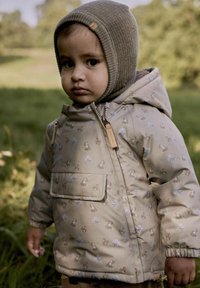 Béžová vzorovaná bunda s kapucňou, obsahujúca zvieracie motívy, prednú kapsu a hnedé akcenty. Textúrovaná pletená kapucňa dokončuje outfit.