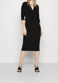 Svart knälång klänning med trekvartslånga ärmar, V-ringning och figurnära design. Matchad med svarta öppetåiga klacksandaler.