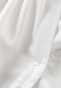 White long-sleeve shirt made of smooth fabric featuring a button detail and subtle textured trim along the placket.