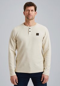 Beige Langarm-Henley-Shirt aus strukturiertem Stoff, mit einem Knopfleiste mit drei Knöpfen und einem kleinen Logo-Patch auf der Brust.