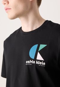 Sort bomuld t-shirt med et farverigt geometrisk logo med blå, grønne og pink former, og hvid tekst "calvin klein" nedenfor.