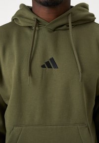 Sweatshirt verde-azeitona feito de tecido macio, com um bolso frontal e um logótipo preto da Adidas. Inclui cordões no capuz.