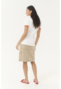 Weißes Poloshirt mit kurzen Ärmeln und orangefarbigen Akzenten, kombiniert mit einem khakifarbenen, knielangen Jeansrock mit Cargo-Taschen und flachen roten Sandalen.