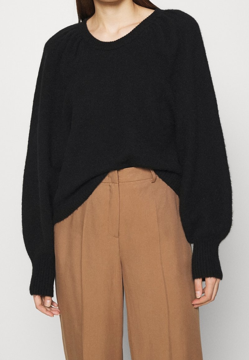 Donna che indossa un maglione nero a maniche lunghe con maniche a palloncino, infilato in pantaloni larghi a vita alta color cammello.