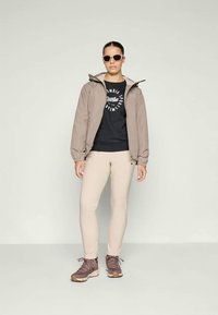 Beige jasje, zwarte sweatshirt met logo, beige leggings en bruine wandelschoenen. Accessoires omvatten zonnebril. Eenvoudige, functionele kleding voor buiten.