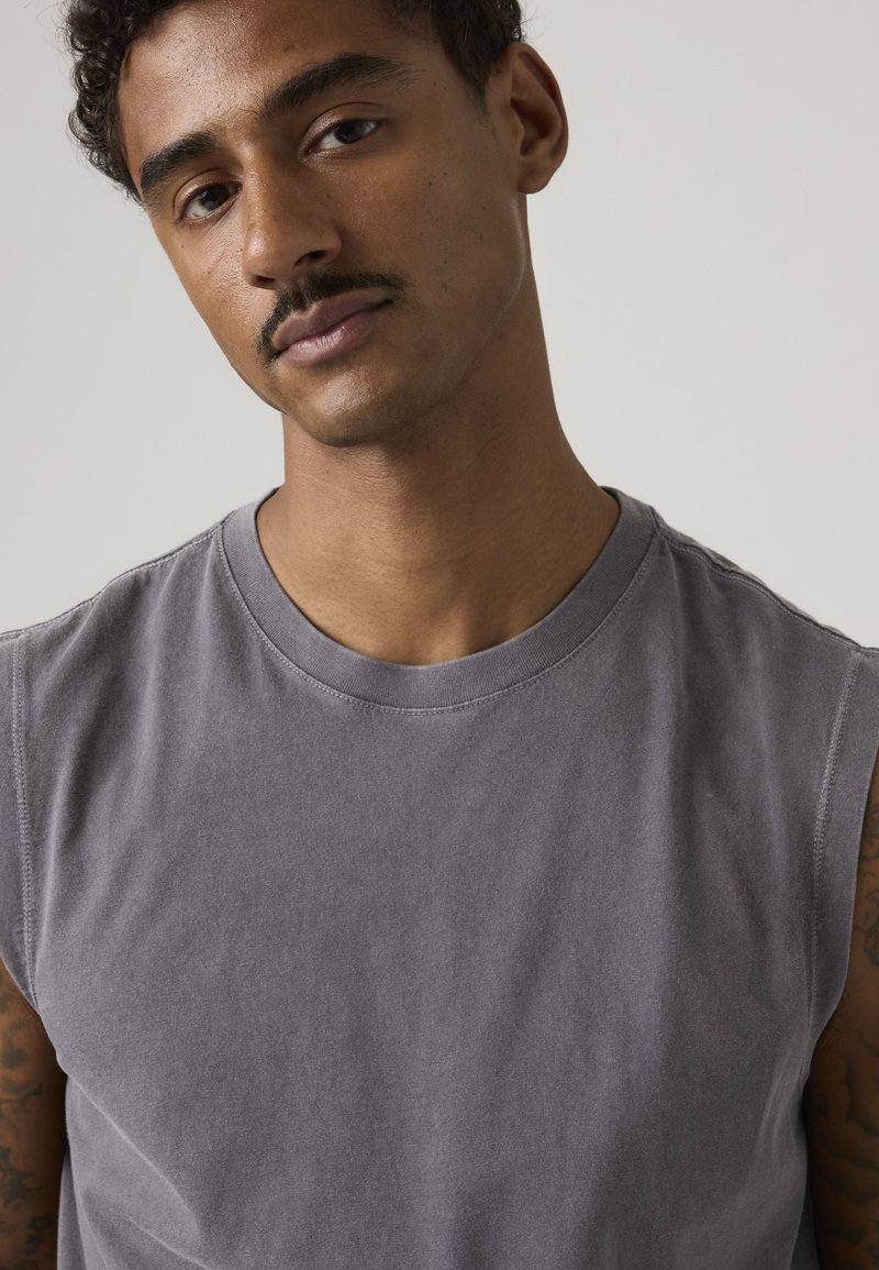 Jeune homme aux cheveux courts et bouclés avec une moustache, portant un débardeur gris, sur un fond clair uni.