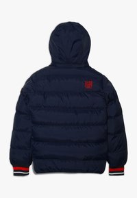 Chaqueta acolchada azul marino con capucha. Cuenta con un logo bordado en rojo en la parte trasera, puños acanalados con franjas rojas, blancas y negras, y un diseño acolchado.