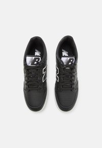 Adidași negri din material textil cu partea superioară din piele, accente albe, cutie perforată pentru degete și șireturi negre. Au un logo vizibil pe limbă și pe branț.