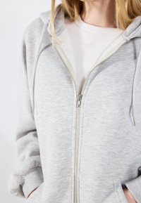 Felpa con zip grigia chiara, dal texture morbido, dotata di una chiusura frontale con cerniera, due tasche laterali e una vestibilità comoda. Indossato sopra un top a coste leggero.