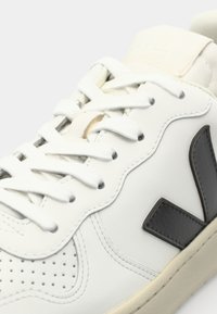 White leather sneakers with perforated detailing, flat laces, and a textured black logo on the side. Features a beige accent on the tongue.