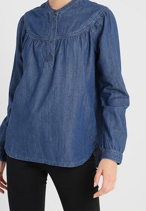 Vrouw draagt een lange mouwen blauwe denim blouse met knoopsluiting, gecombineerd met zwarte broek, staand tegen een effen achtergrond.