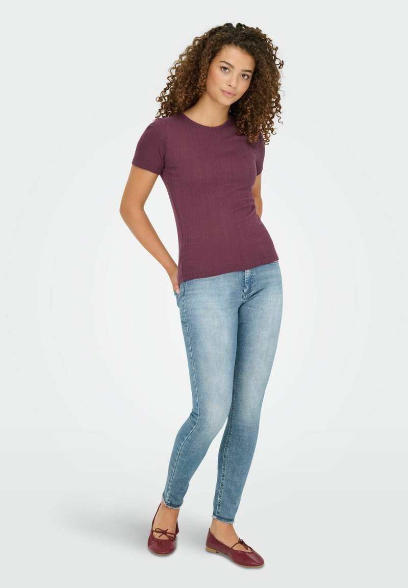 Burgunderfarbens, strukturiertes Kurzarmoberteil, kombiniert mit hellblauen, enganliegenden Jeans und maronenfarbigen Ballerinas mit einer Schleifendetail.