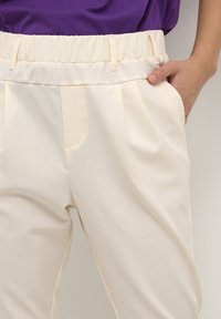 Person med lysebrune bukser med elastisk talje og bæltestropper, hånden hvilende i den højre lomme, lilla skjorte synlig ovenfor.