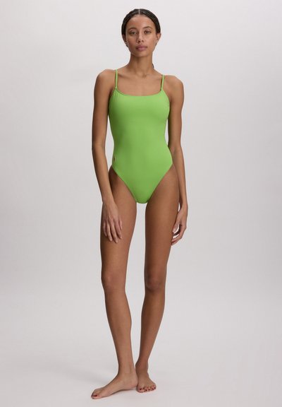 Γυναίκα που στέκεται ξυπόλητη φορώντας στενή πράσινη ολόσωμη φόρμα με λεπτά ιμάντες ώμων σε ουδέτερο φόντο.