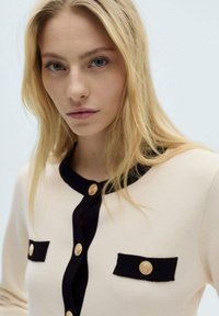 Beige knit cardigan with black accents, featuring a round neck, gold buttons, and two front pockets with ribbed detailing.