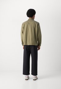 Khaki button-up jacket with a relaxed fit, long sleeves, and a small collar. Paired with black wide-leg pants and shoes with three stripes.