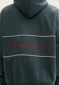 Ciemnozielona bluza z kapturem, z czerwonym haftem z napisem "Academy Club" i białym paskiem na plecach. Wykonana z materiału bawełnianego.