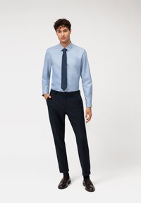 Himmelblaues Langarmhemd mit Button-Down-Kragen, kombiniert mit dunkelblauen Hosen und dunkelbraunen Slippern, vor einem neutralen Hintergrund präsentiert.