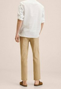Person in einem weißen Langarmshirt, beigen Hosen und braunen Slippern, steht vor einem schlichten hellen Hintergrund.