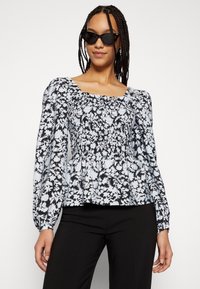Blusa floral com decote quadrado, corpete pregueado e mangas bufantes longas em preto e azul claro. Combinada com calças pretas.