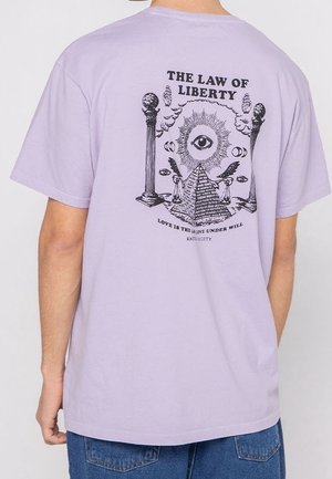 Osoba w lawendowej koszulce z grafiką na plecach przedstawiającą oko, piramidę, kolumny, skrzydlate postacie oraz napis „The Law of Liberty”.