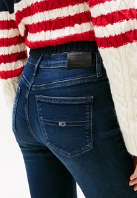 Asmuo dėvi tamsiai mėlynas džinsines kelnes su mažu logotipu užnugario kišenėje ir raudonai, baltai bei tamsiai mėlynai megztą megztinį su pynimu.