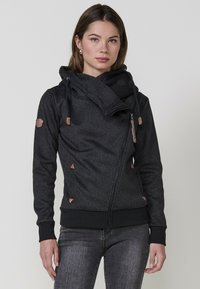 Felpa nera con zip e colletto alto, tessuto testurizzato, dettagli triangolari e particolari simili alla pelle. Presenta un cordino regolabile.