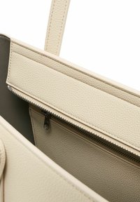 Interno di una borsa a mano in pelle beige con una tasca con zip, superficie testurizzata e finitura liscia. Presenta due manici e un ampio scomparto principale.