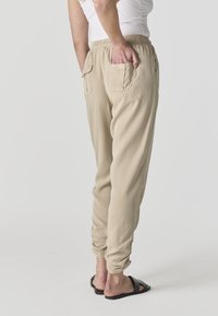 Personne portant un pantalon beige à taille élastique avec des poches arrière et des sandales slides noires, une main placée dans une poche, debout sur un fond gris.