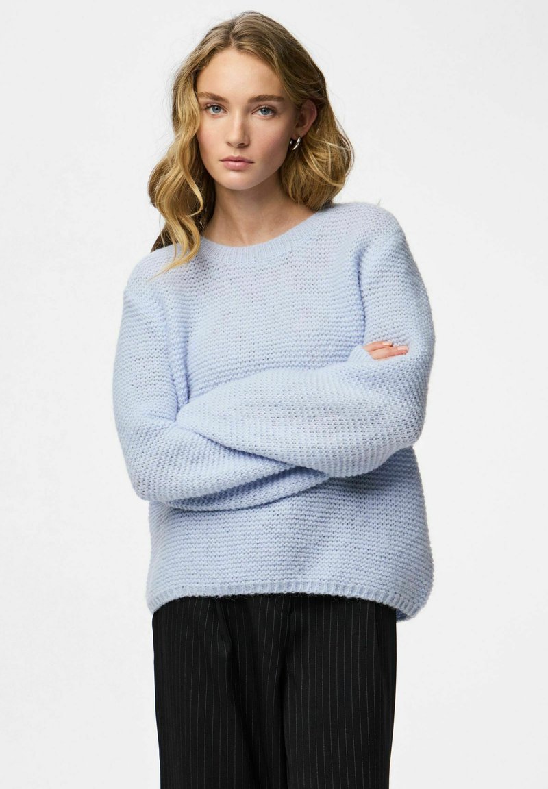 Pull tricoté bleu clair avec un col rond, coupe décontractée et poignets côtelés. Porté avec un pantalon noir à fines rayures. Texture douce.
